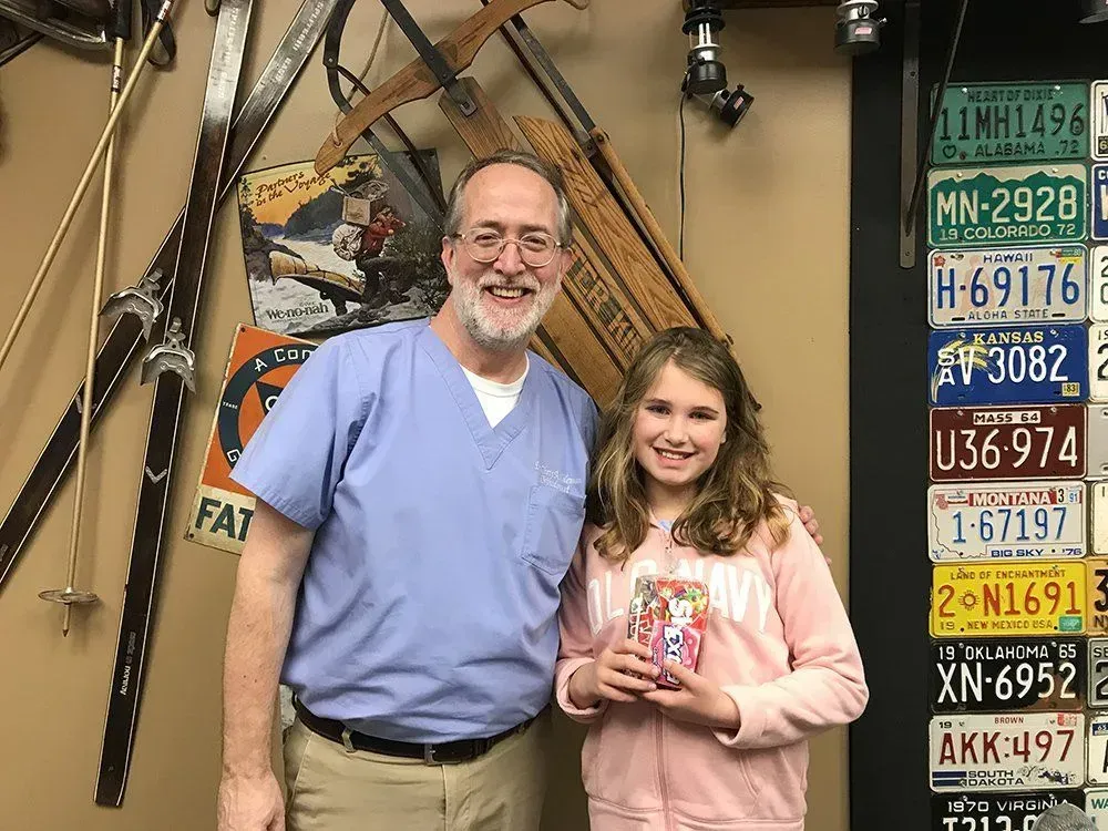 Man in scrubs and girl smiling, holding a gift, in a room with license plates and swords.