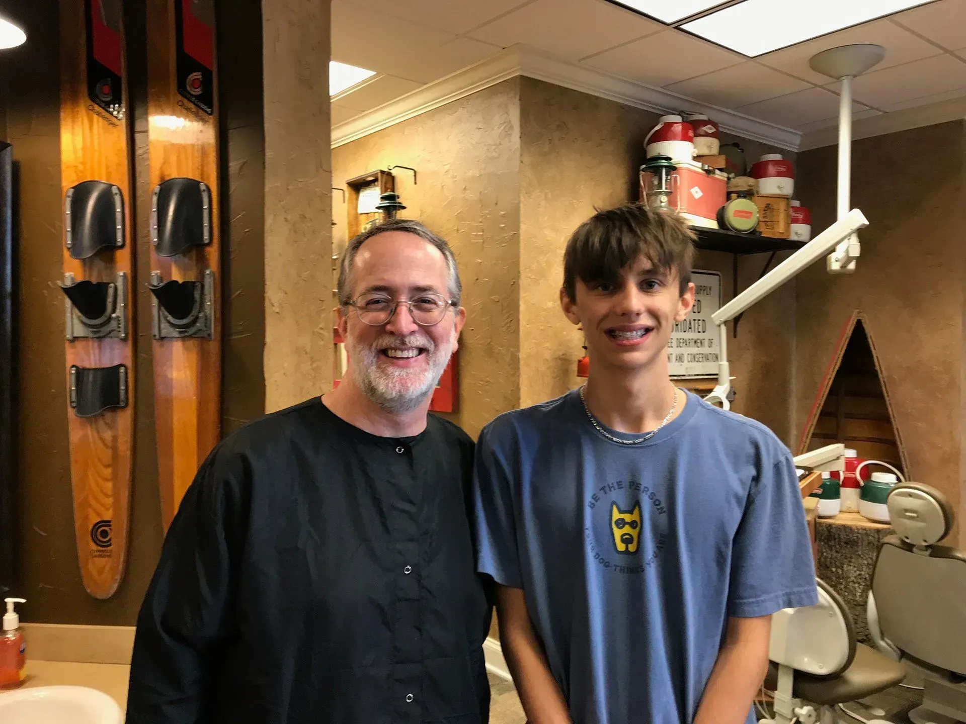 A man in dental scrubs and a teen with braces smiling in a dental office. Water skis on wall.