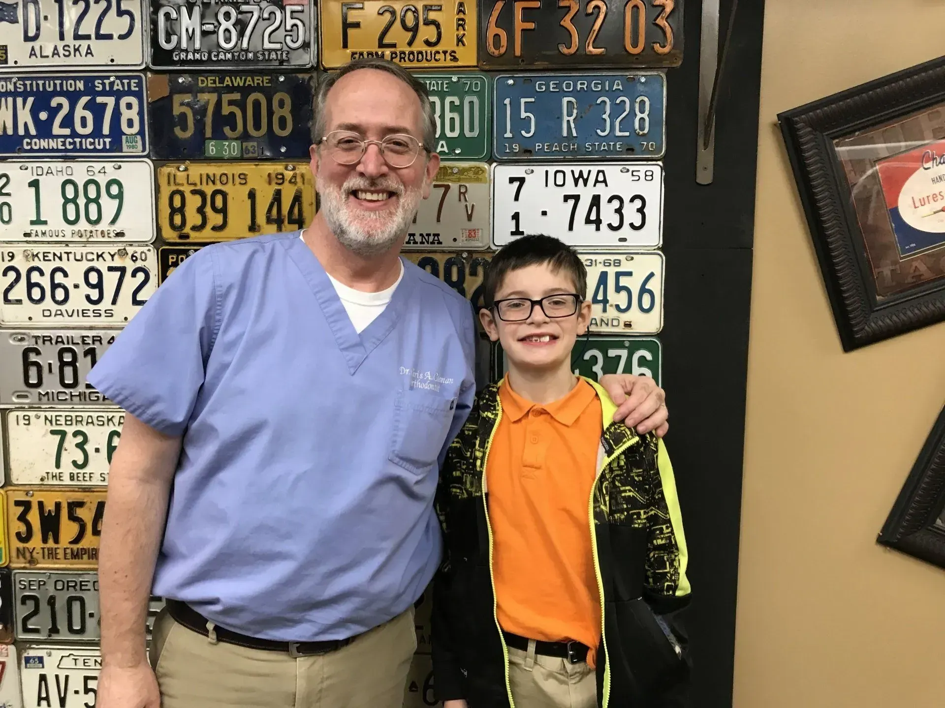 A man in blue scrubs and a boy in a yellow shirt stand by a wall of license plates, smiling.