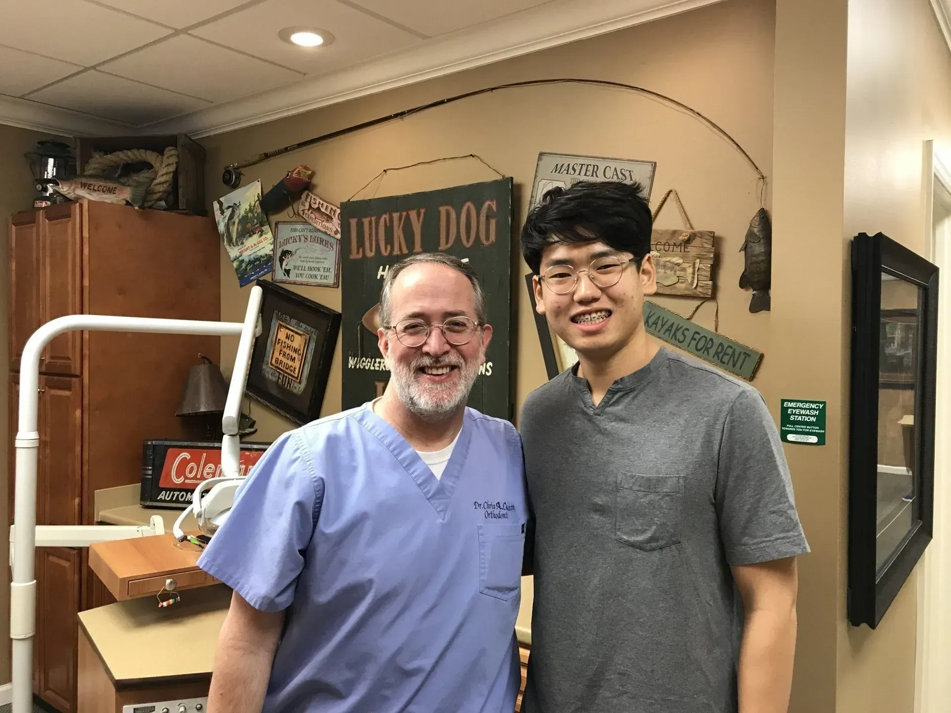 Two men smiling in a dental office. One in scrubs, the other in a gray t-shirt. Beige walls with decor.