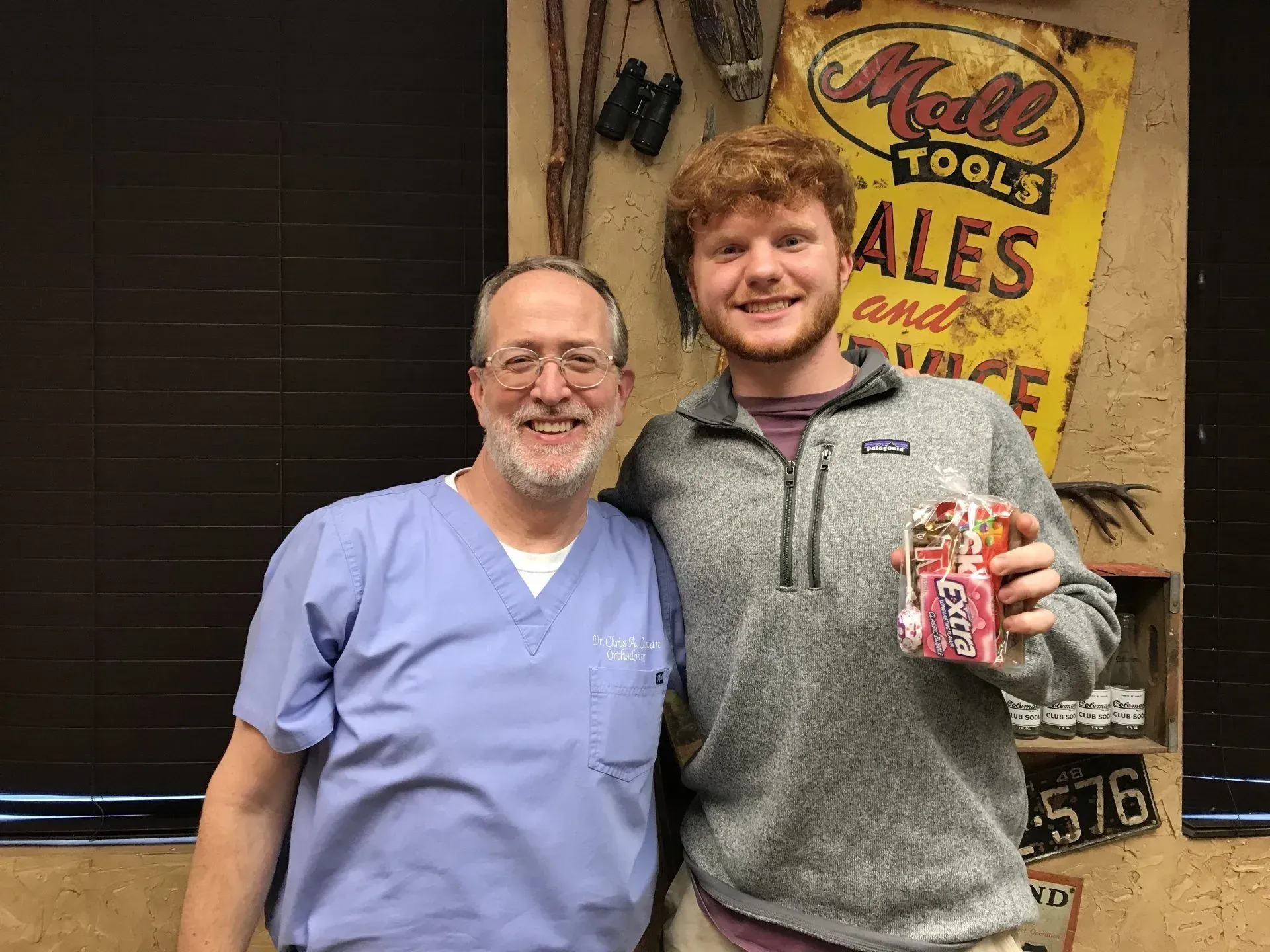 Two men smile; one in scrubs, one holding candy. 