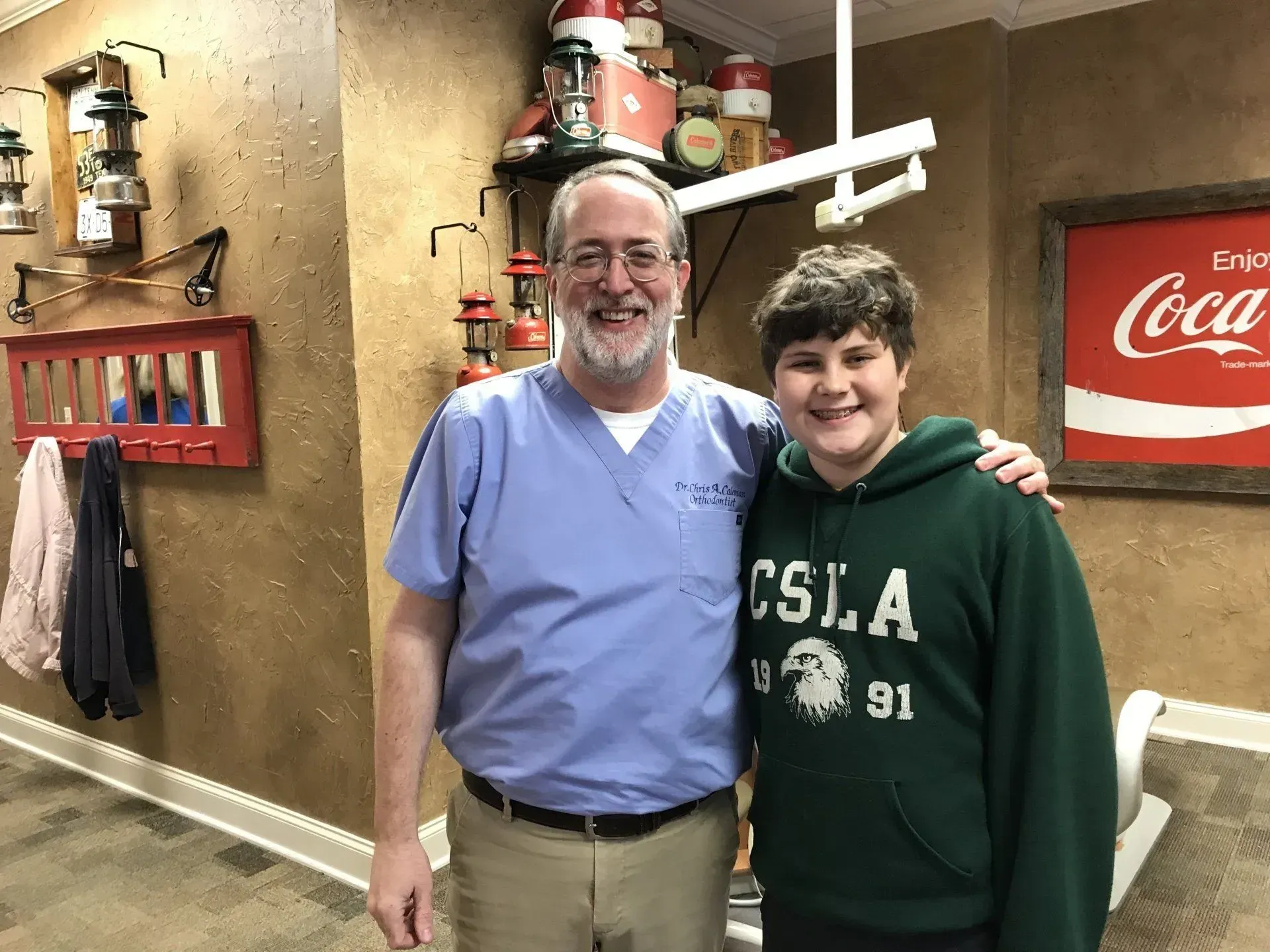 Man in scrubs and boy in green hoodie smiling, pose by a Coca-Cola sign in a room with vintage decor.