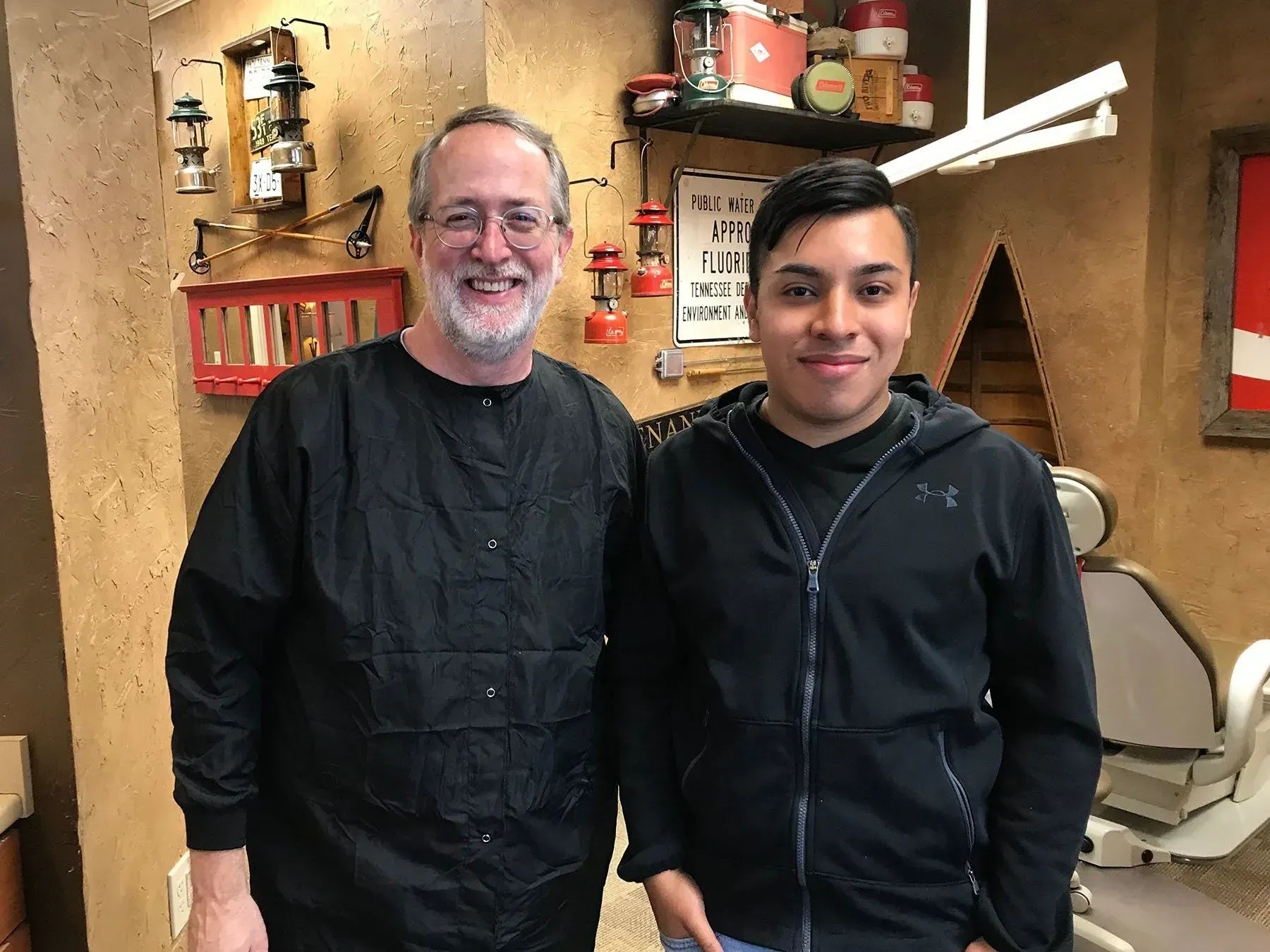 Man with glasses and another man in a black jacket smiling in a dental office.