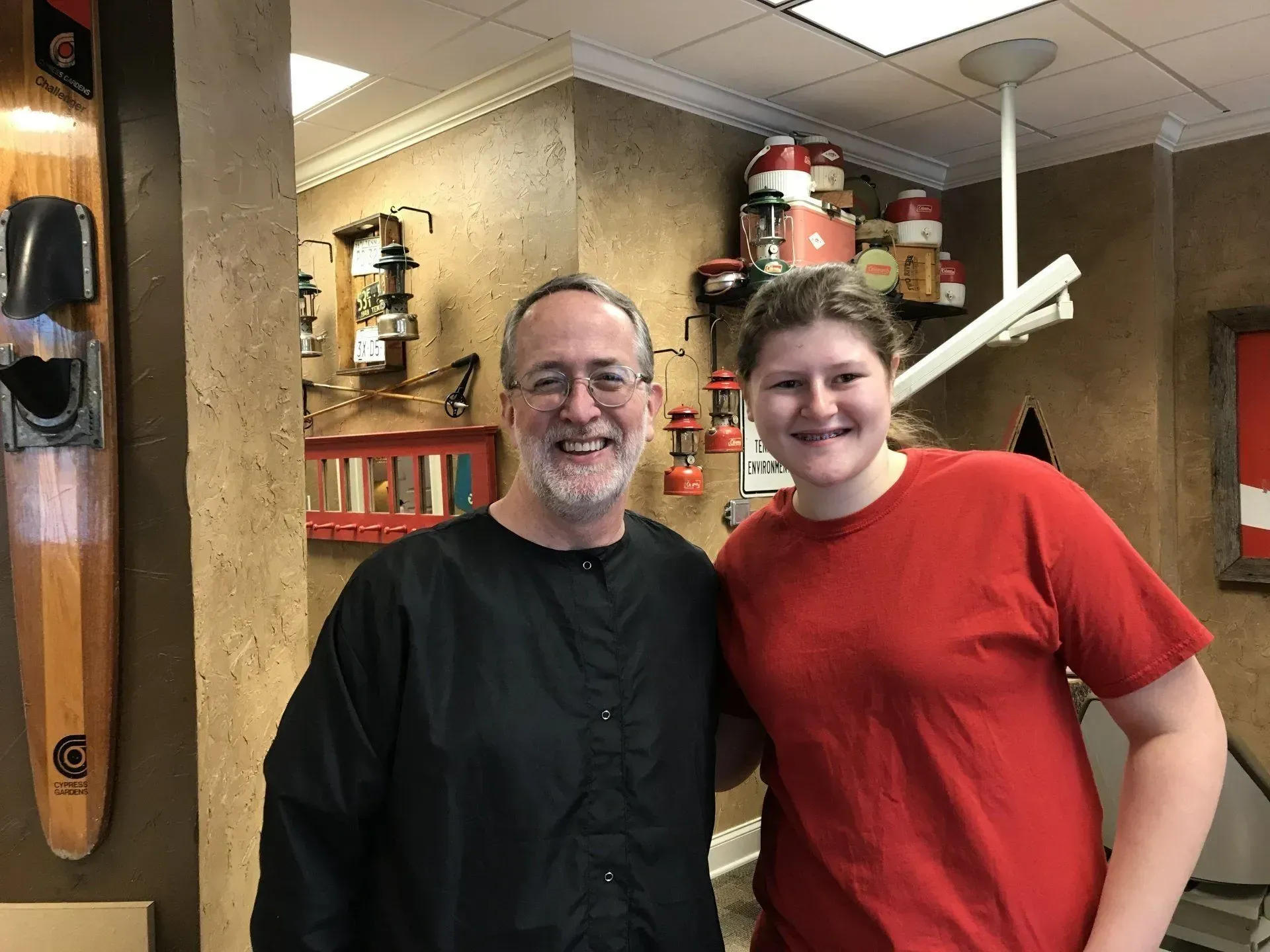 Man and young person smiling and posing together inside a dental office. The man wears black, the young person wears red.