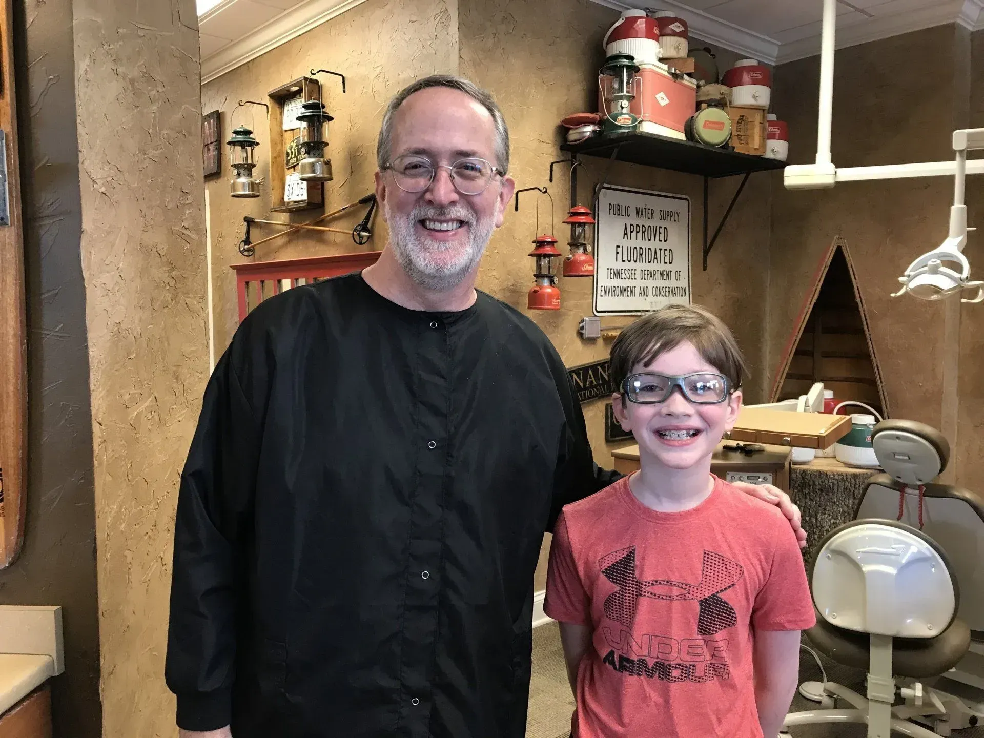 Dentist with a young boy in a dental office. Both are smiling, boy is wearing glasses. Rustic décor.
