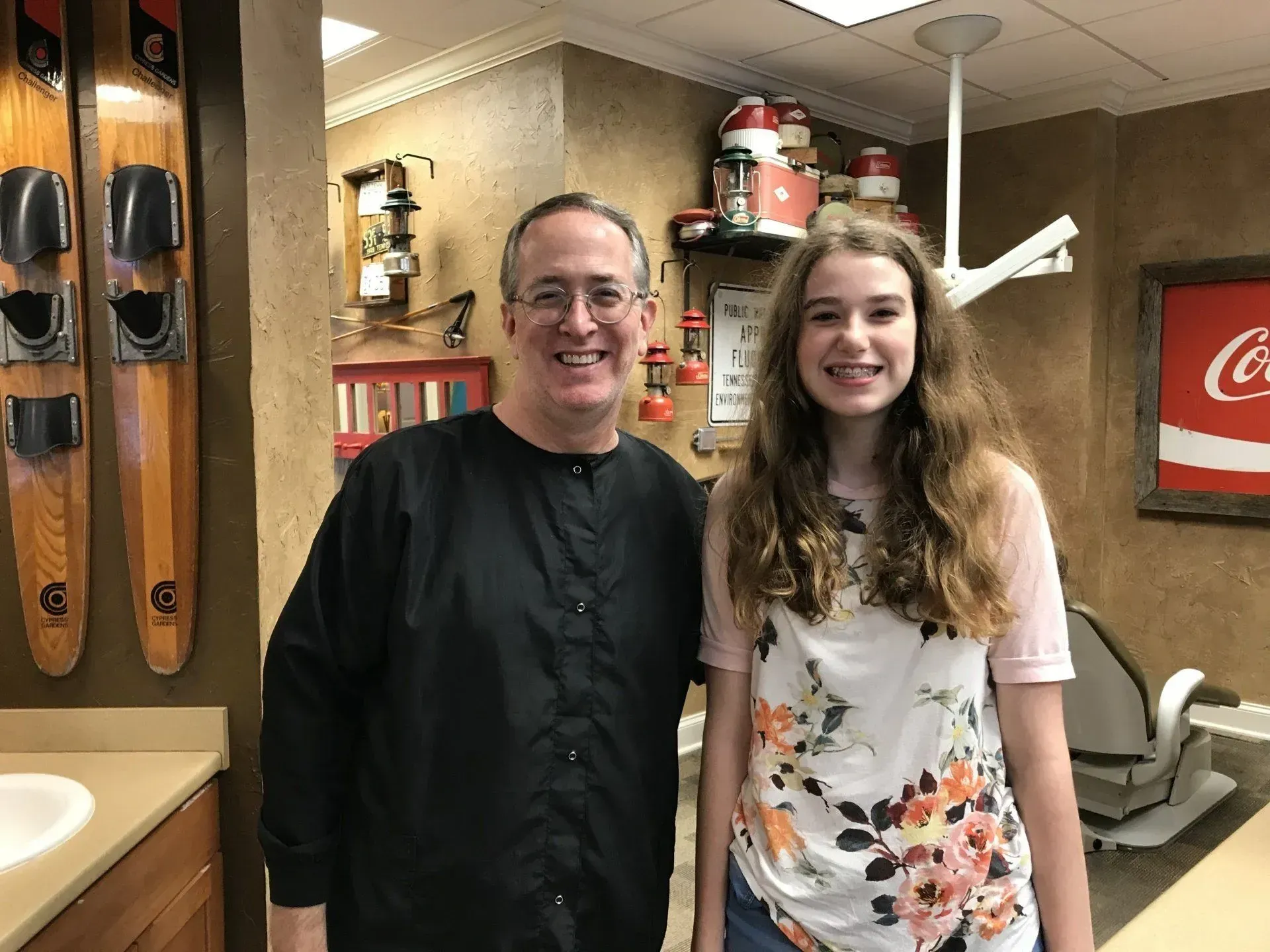 Man and girl smile together in a dental office. The girl has braces. Office decorated with water skis and Coca-Cola sign.