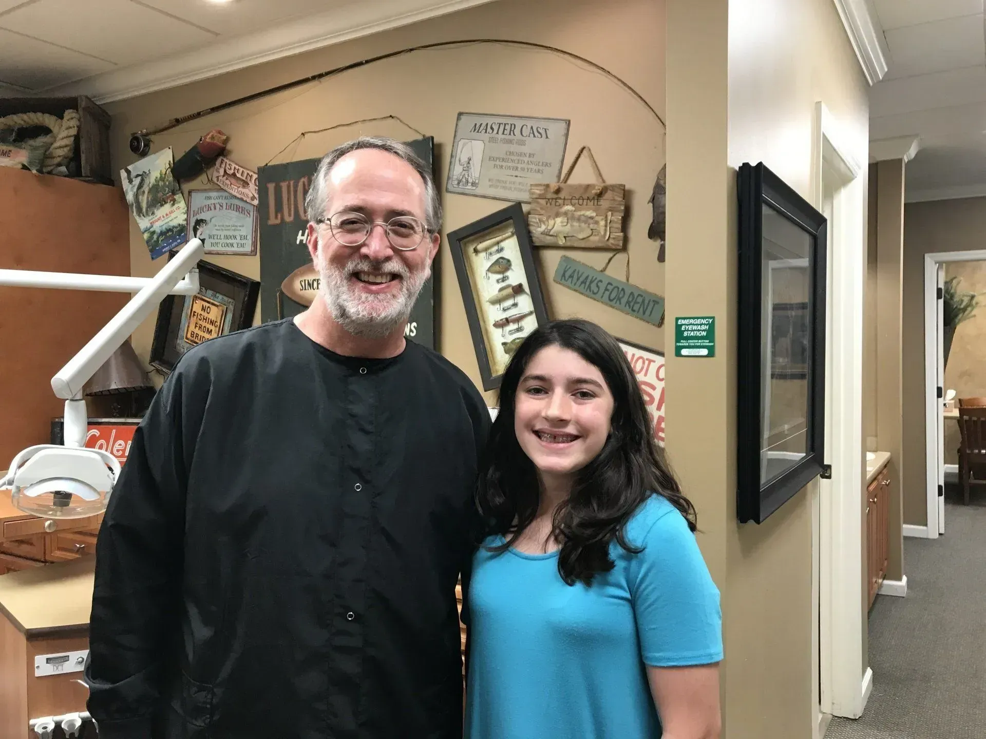 A smiling dentist with a young person in a dental office. The person is wearing a teal shirt and has braces.