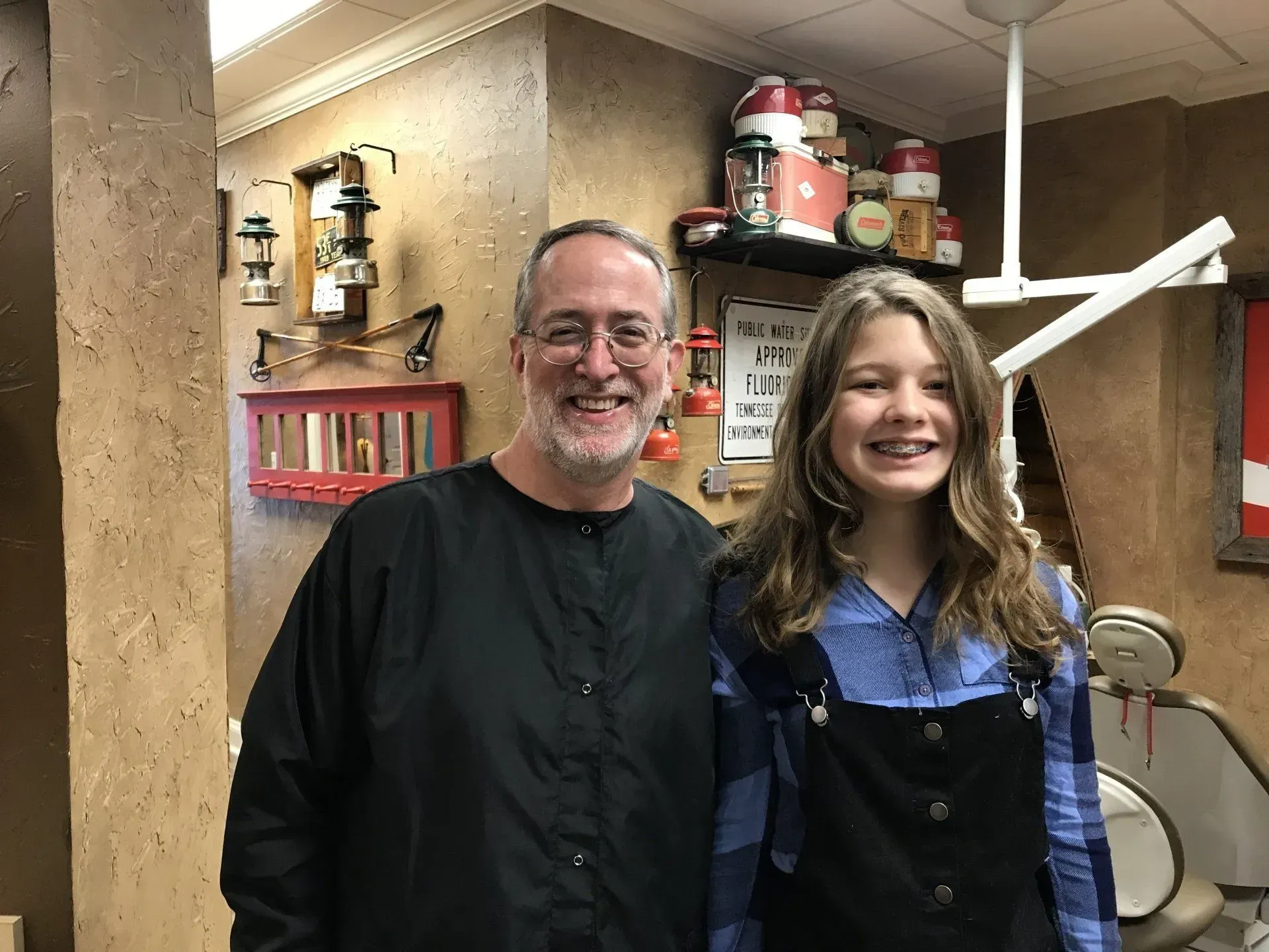A smiling person with glasses and a girl with braces pose in a dental office.