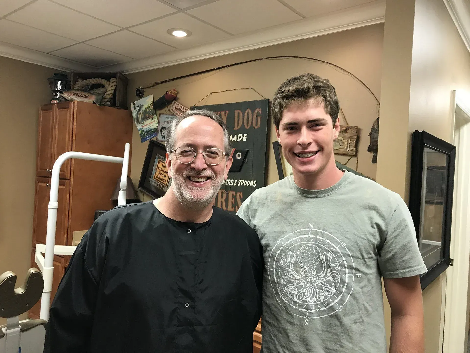 Man in black cape with a smiling young man, pose together in a barbershop.