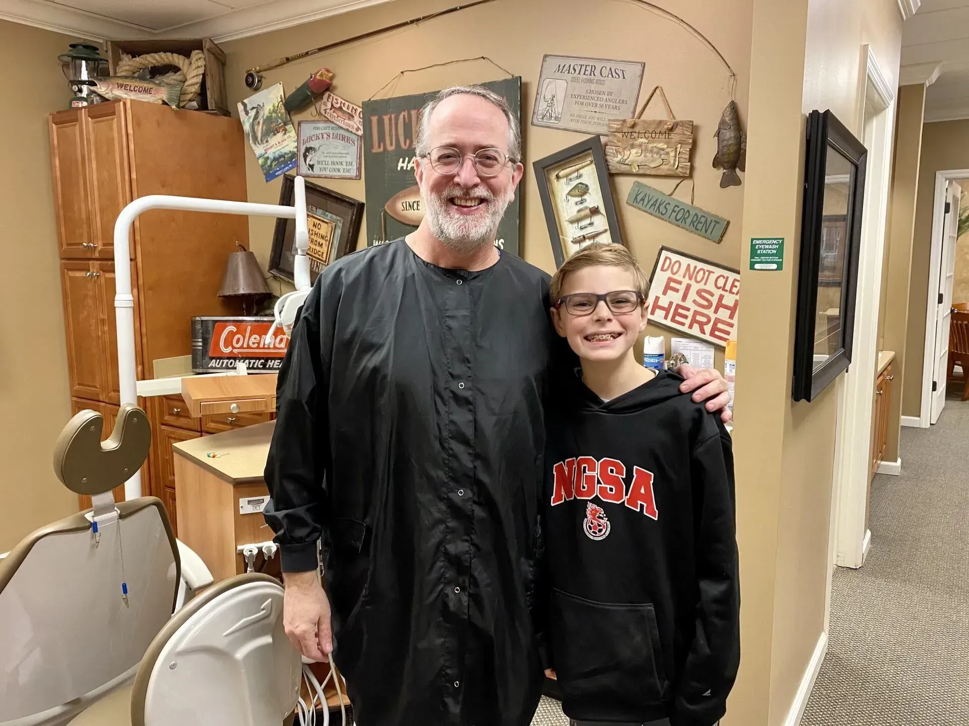 Man in black smock and boy in hoodie pose in a dental office. Both smile, arm around the boy. Fishing theme decor.