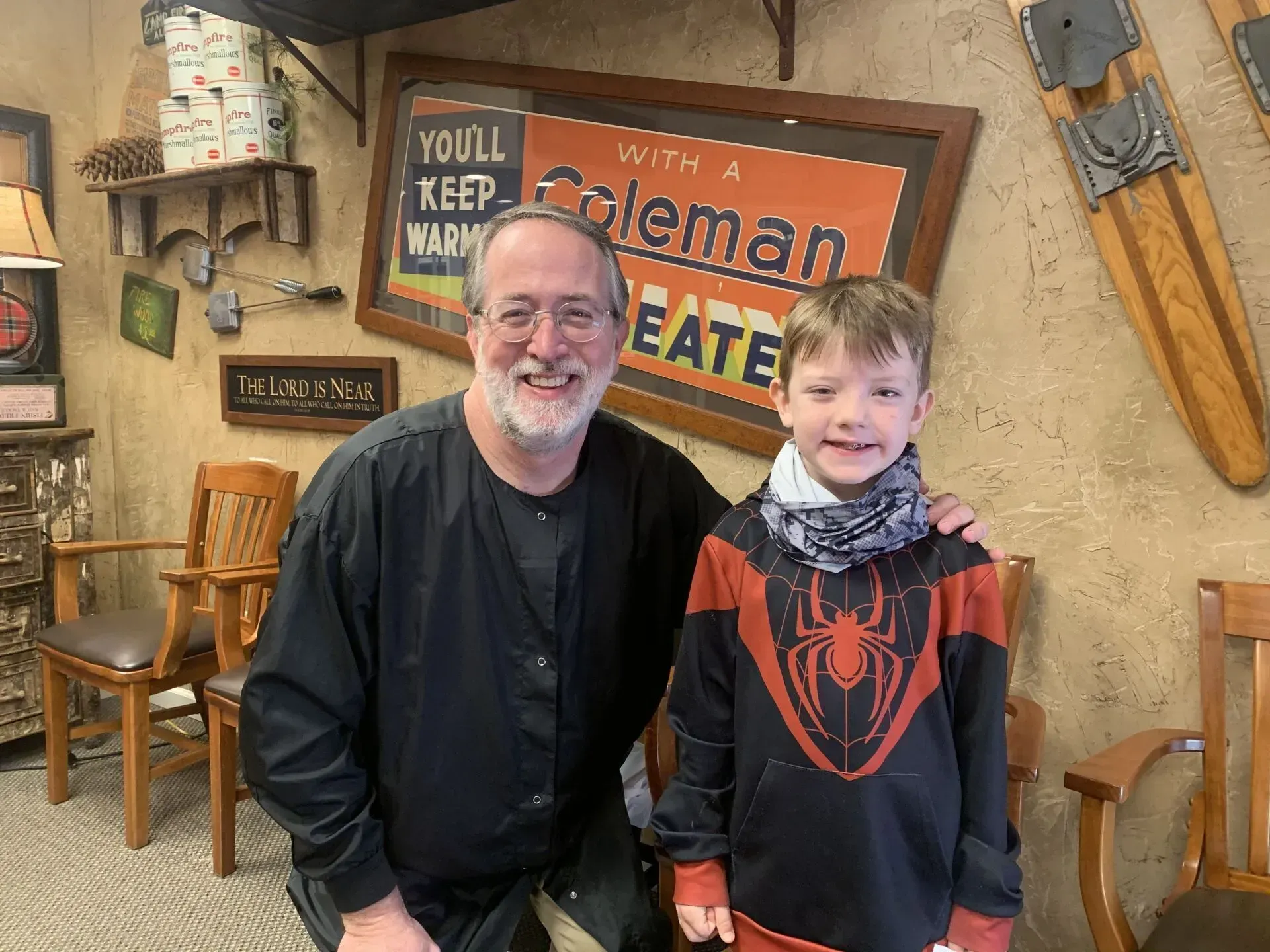 Man and boy smiling together, pose in room with antique decor. Man in black scrubs, boy in Spider-Man hoodie.
