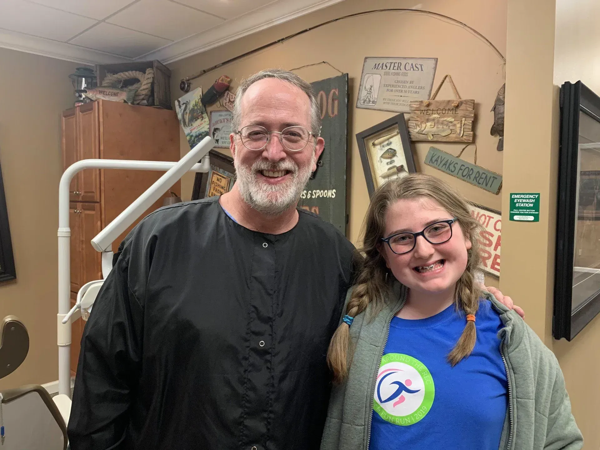 Dentist and patient smiling together in dental office. Man in black smock, girl in blue shirt.