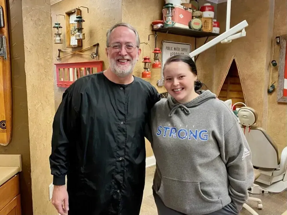 Dentist and patient smiling together in a rustic-themed dental office. The patient wears a gray sweatshirt.