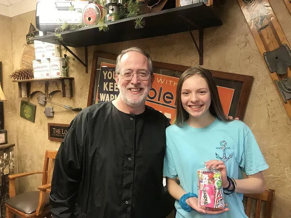 A man and a girl smiling, holding a gift, in a salon. The man wears a black smock.
