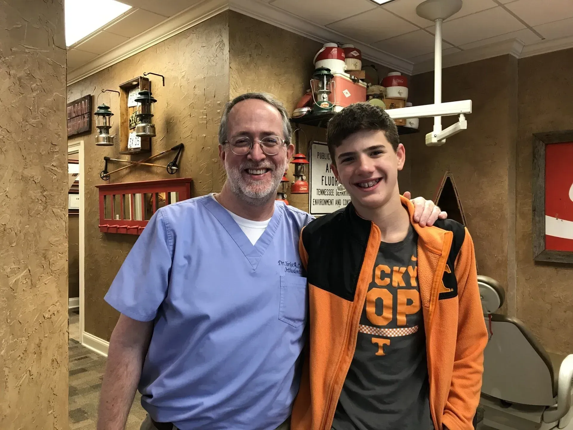 Man in scrubs and a teen with braces pose together in a dental office. The teen wears orange.
