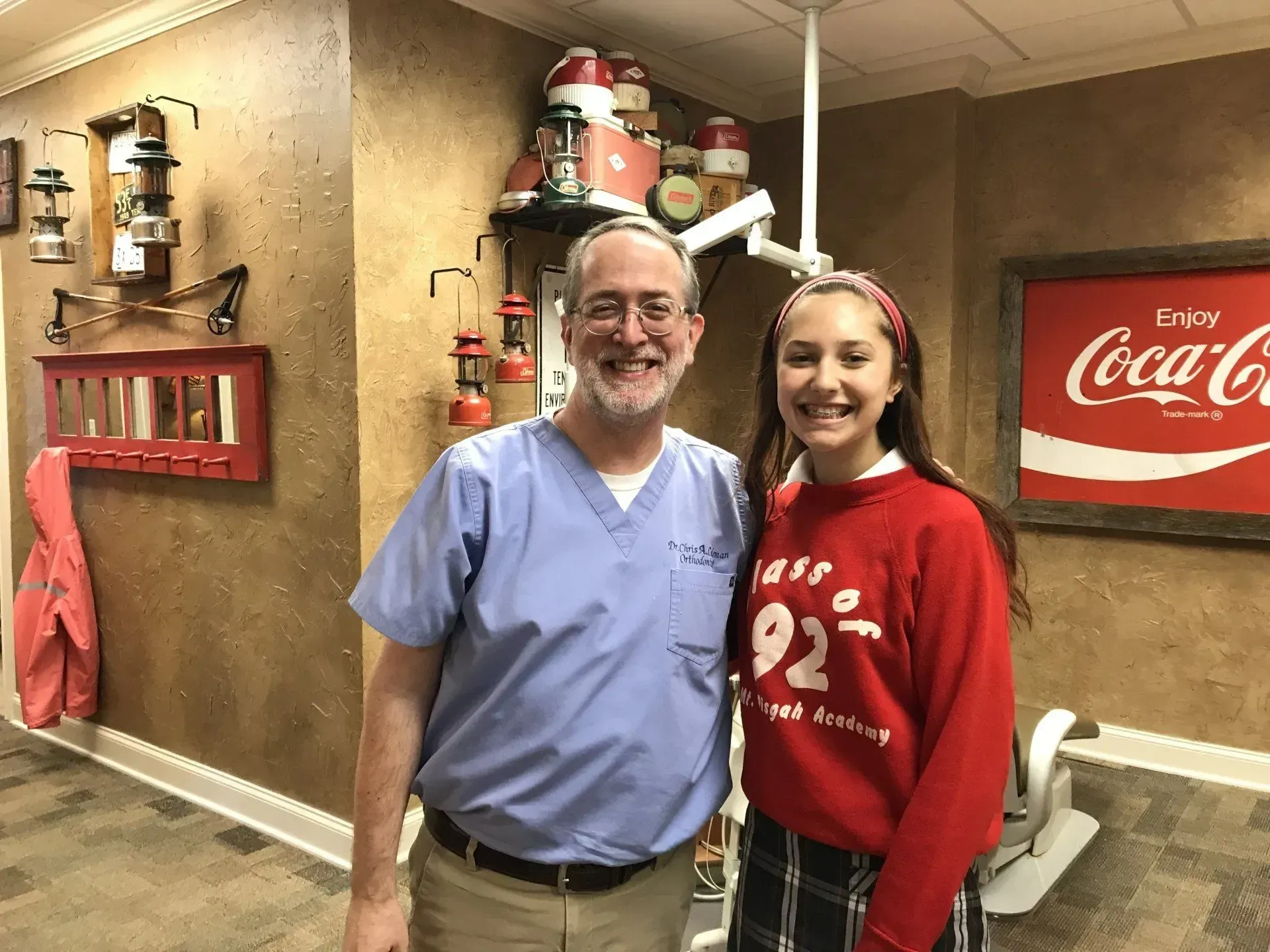 Man in scrubs and young person smile, next to Coca-Cola sign in room with vintage decor.