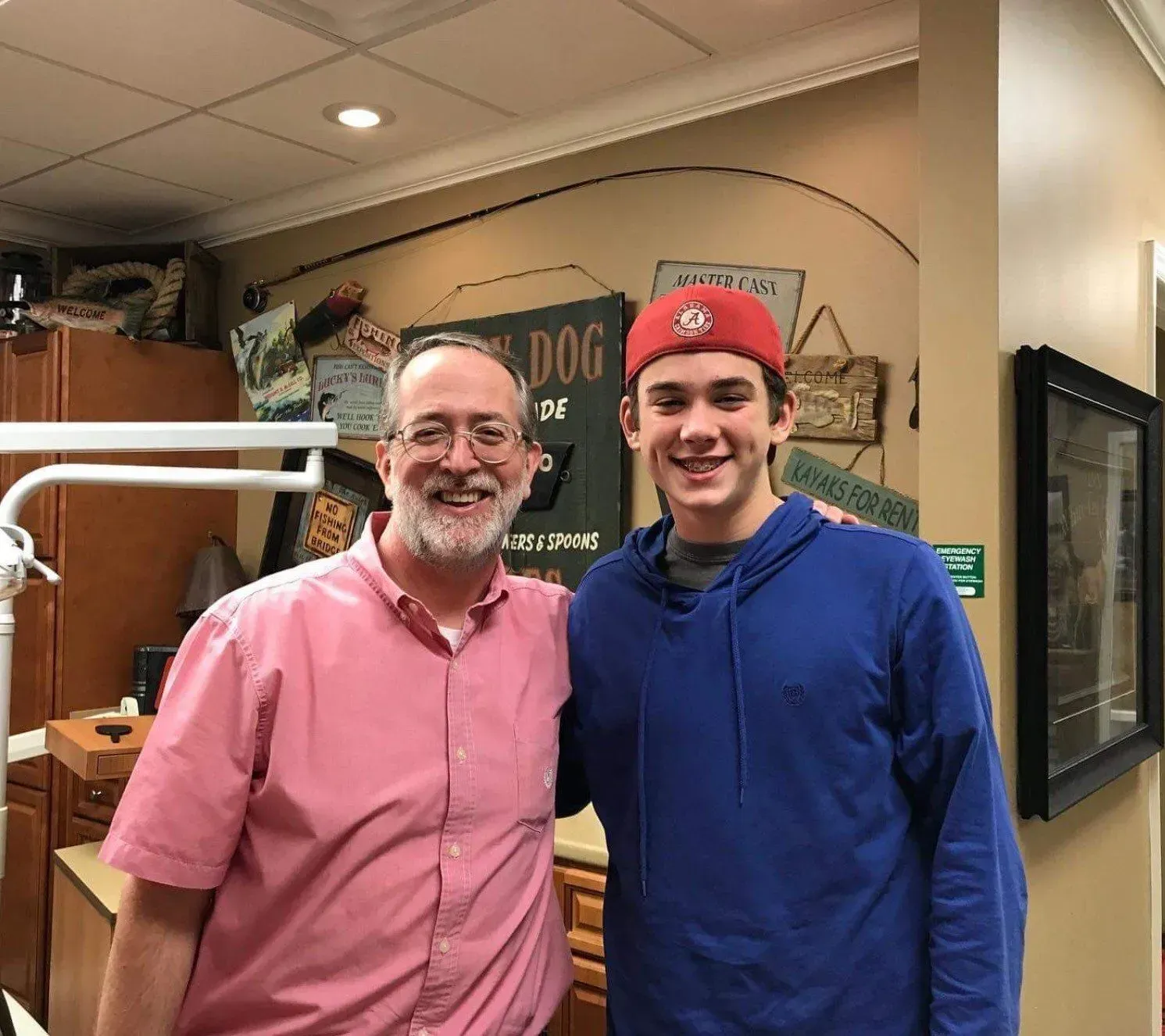 Man in pink shirt and teen in blue hoodie pose inside a dental office, smiling.
