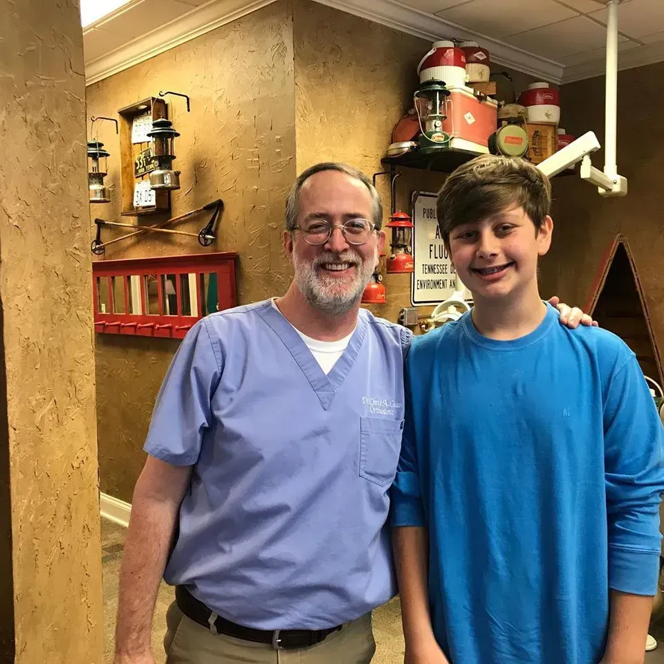 Man in blue scrubs with arm around a teen in blue shirt, smiling in a dental office.