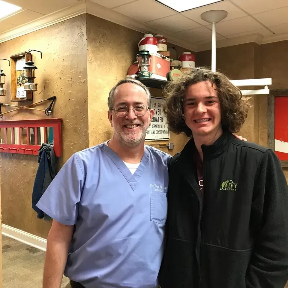 Man in scrubs and young person smile, pose inside a business, near decorative shelving and wall art.
