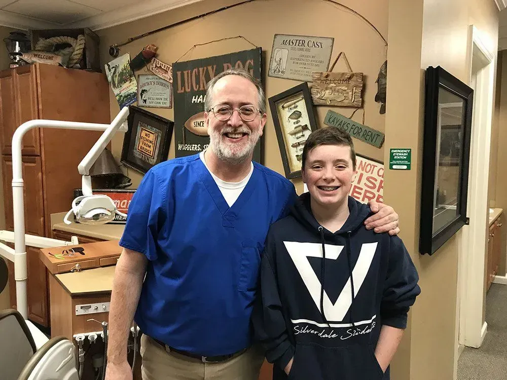 A dentist in blue scrubs poses with a smiling teen in a blue hoodie, arm around his shoulder in a dental office.