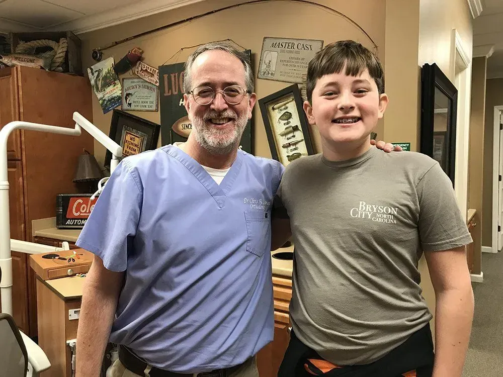 Dentist and young patient smiling, posing in dental office. Dentist in scrubs, arm around patient's shoulder.