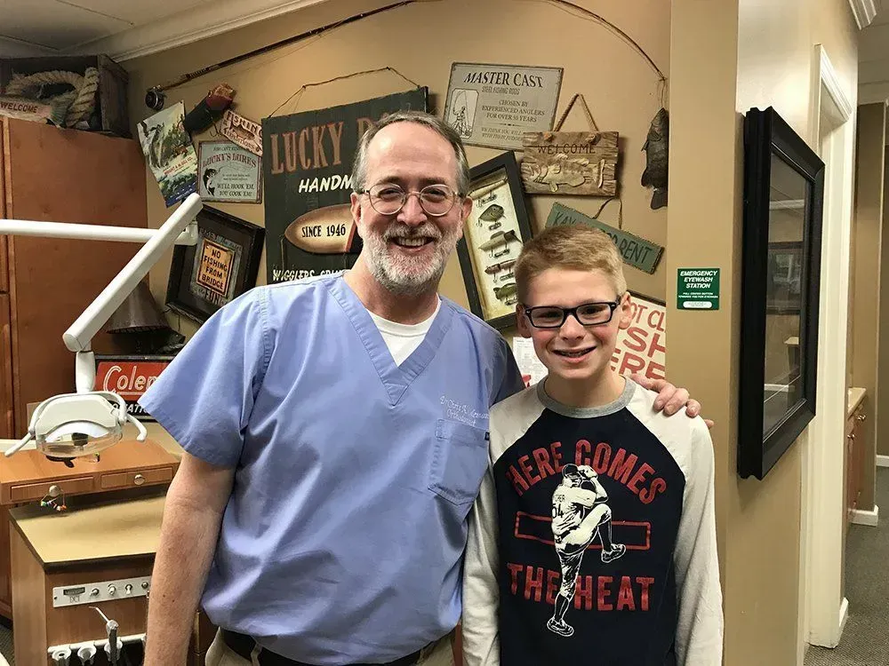 Man in scrubs and boy with glasses pose together in dental office. Smiling.