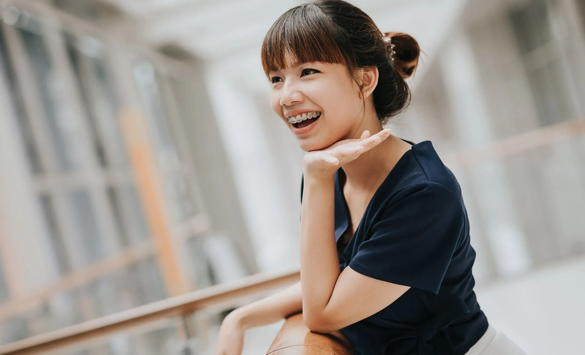 Woman with braces smiles, resting her chin on her hand, in a bright, modern building.