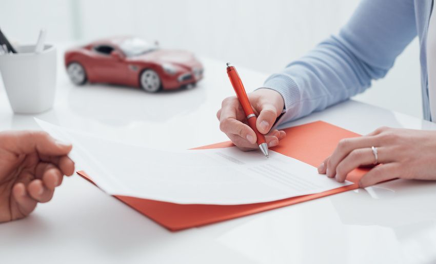 Hands signing a car purchase agreement beside a red model car on a white desk