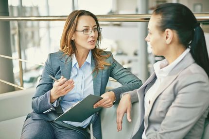 Woman in glasses gestures while speaking to another woman in a business setting.