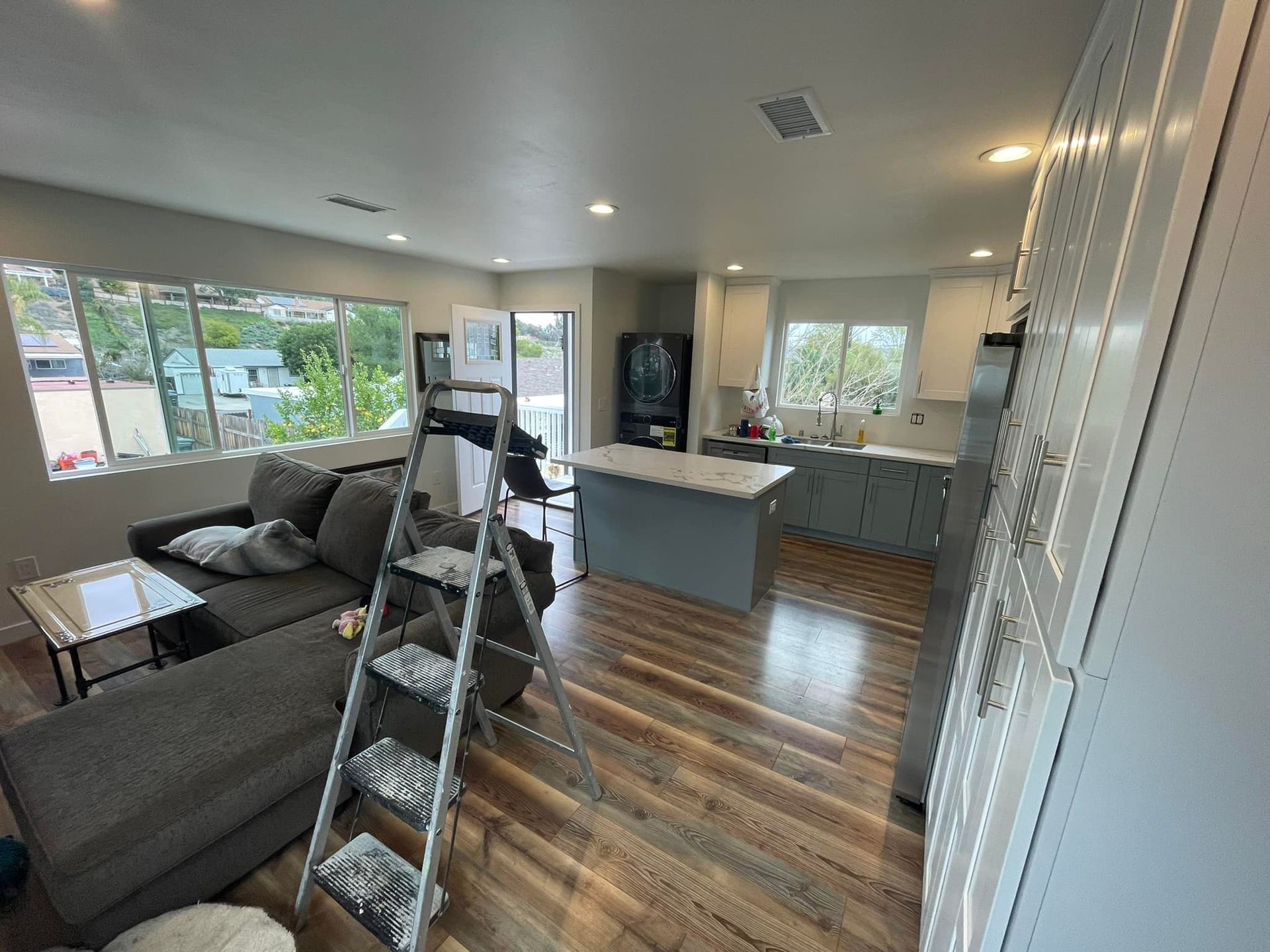 Living room with gray sofa, kitchen island, and a stepladder; hardwood floors and large windows.