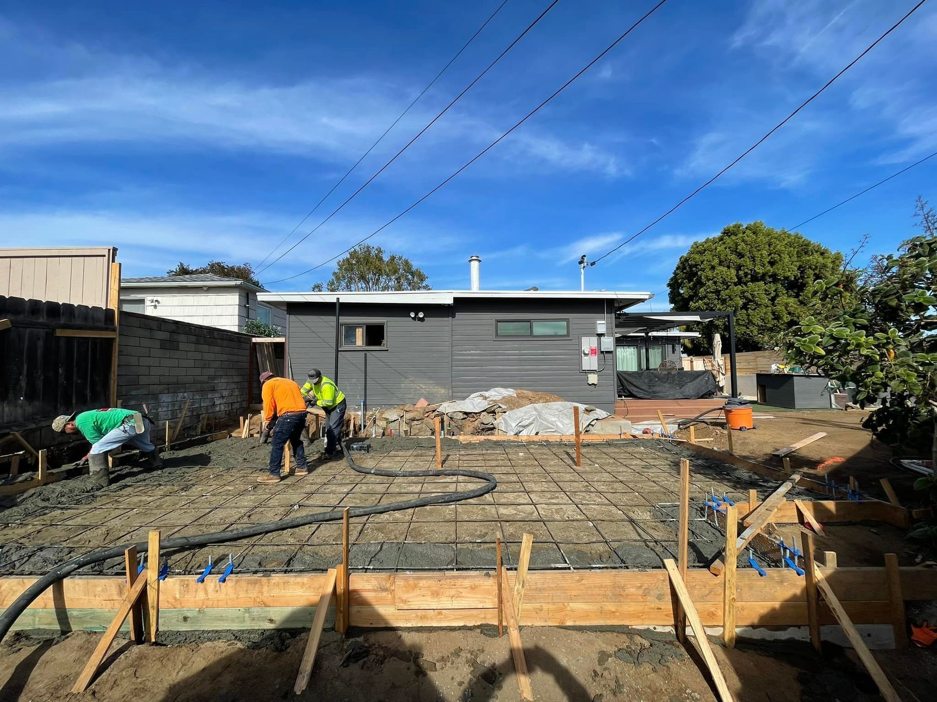 Construction workers pouring concrete for a foundation, wooden forms and rebar visible. Houses and blue sky in background.