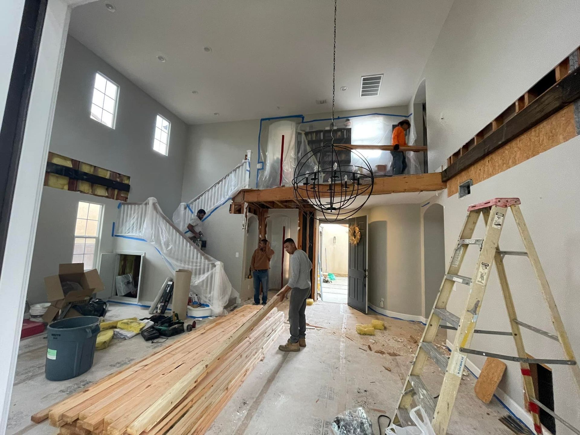 Construction workers inside a home remodel. Staircase, walls and ceiling are partially demoed. Lumber and equipment are present.