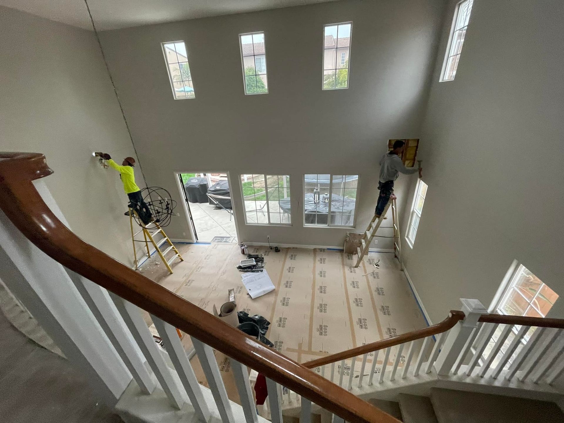 Workers on ladders installing or repairing drywall inside a house with high ceilings and stairs.