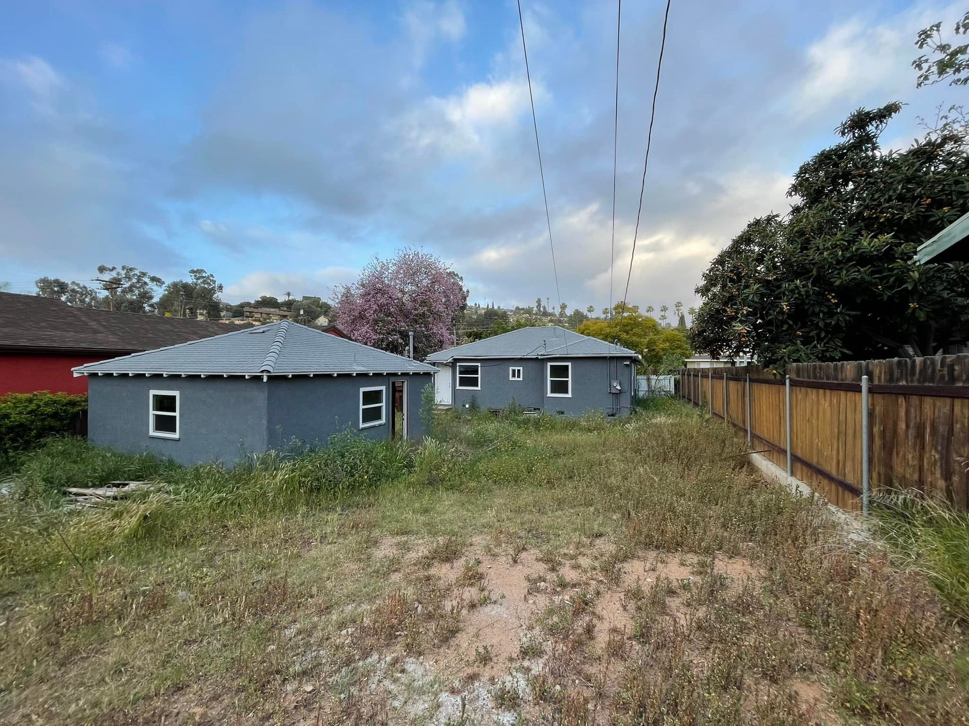 Two small, gray buildings with white-framed windows sit in an overgrown lot, near a wooden fence and flowering tree.