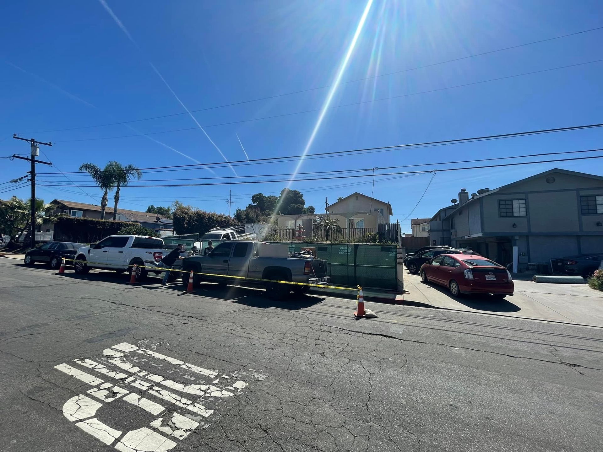 Street scene with a truck hauling a large green dumpster filled with yard waste; houses and parked cars line the street.