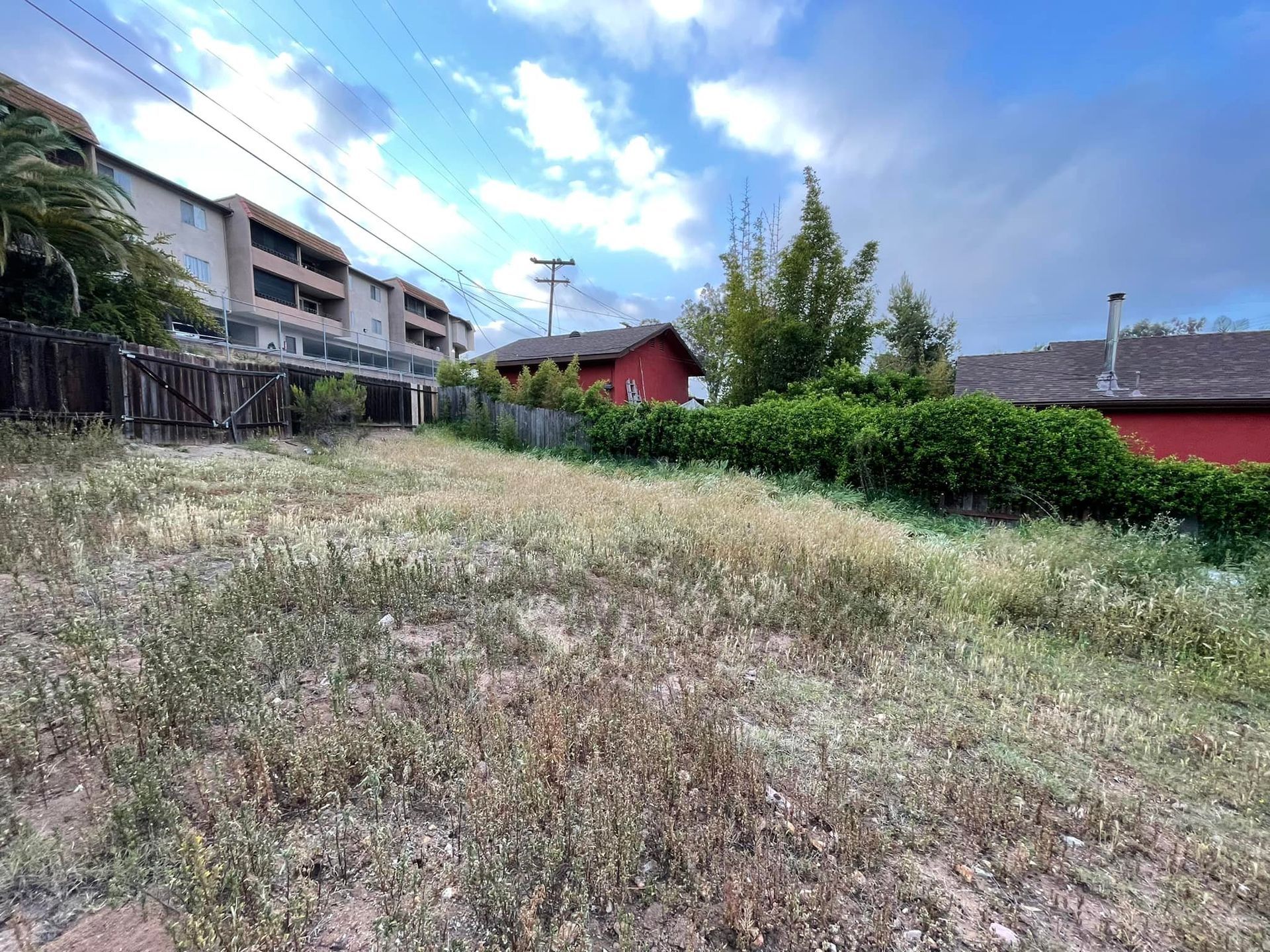 Grassy vacant lot with a wooden fence, red house, and apartment building under a cloudy sky.