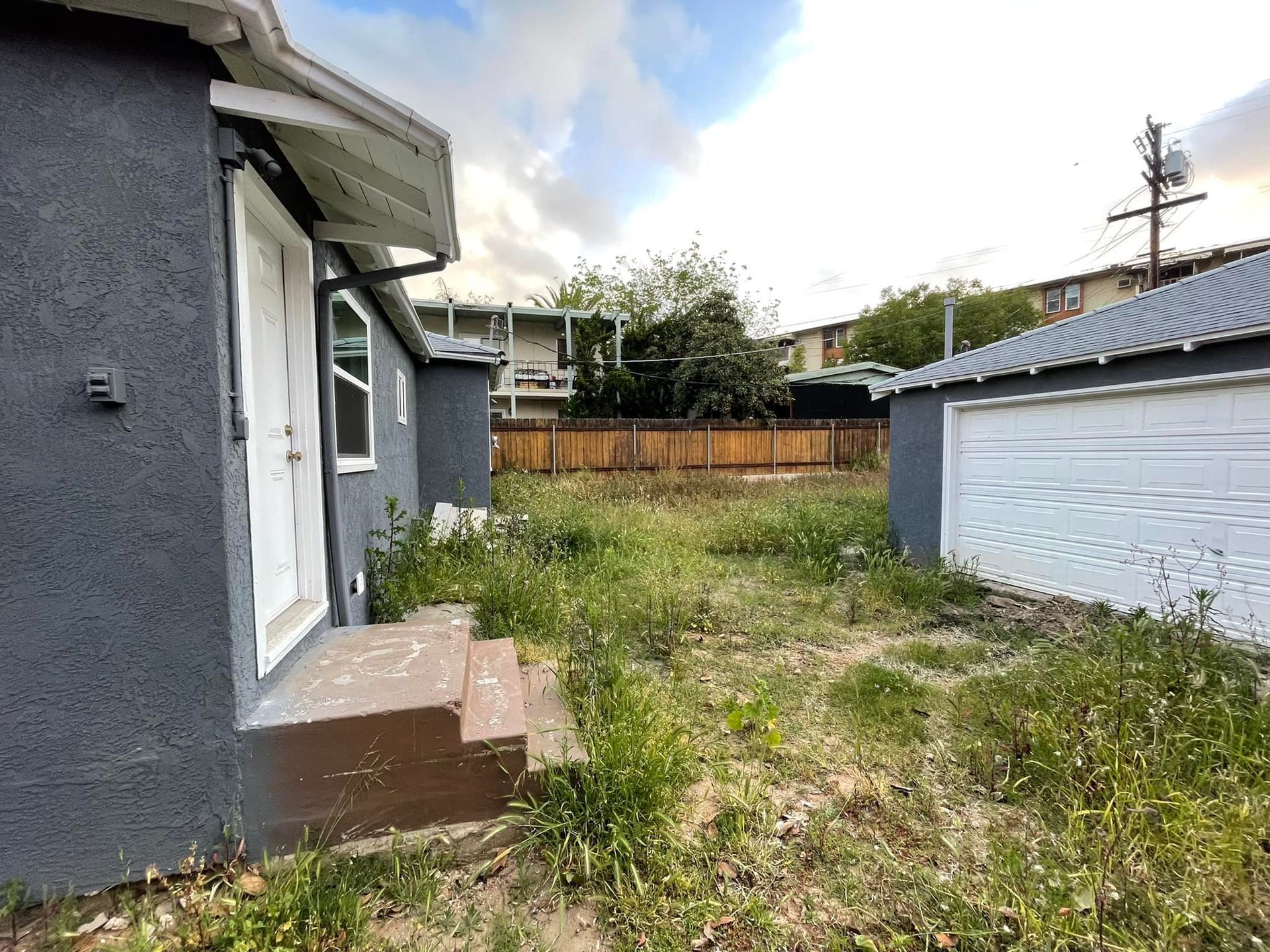 Exterior view of a gray building with a white door and a garage, overgrown yard with weeds.