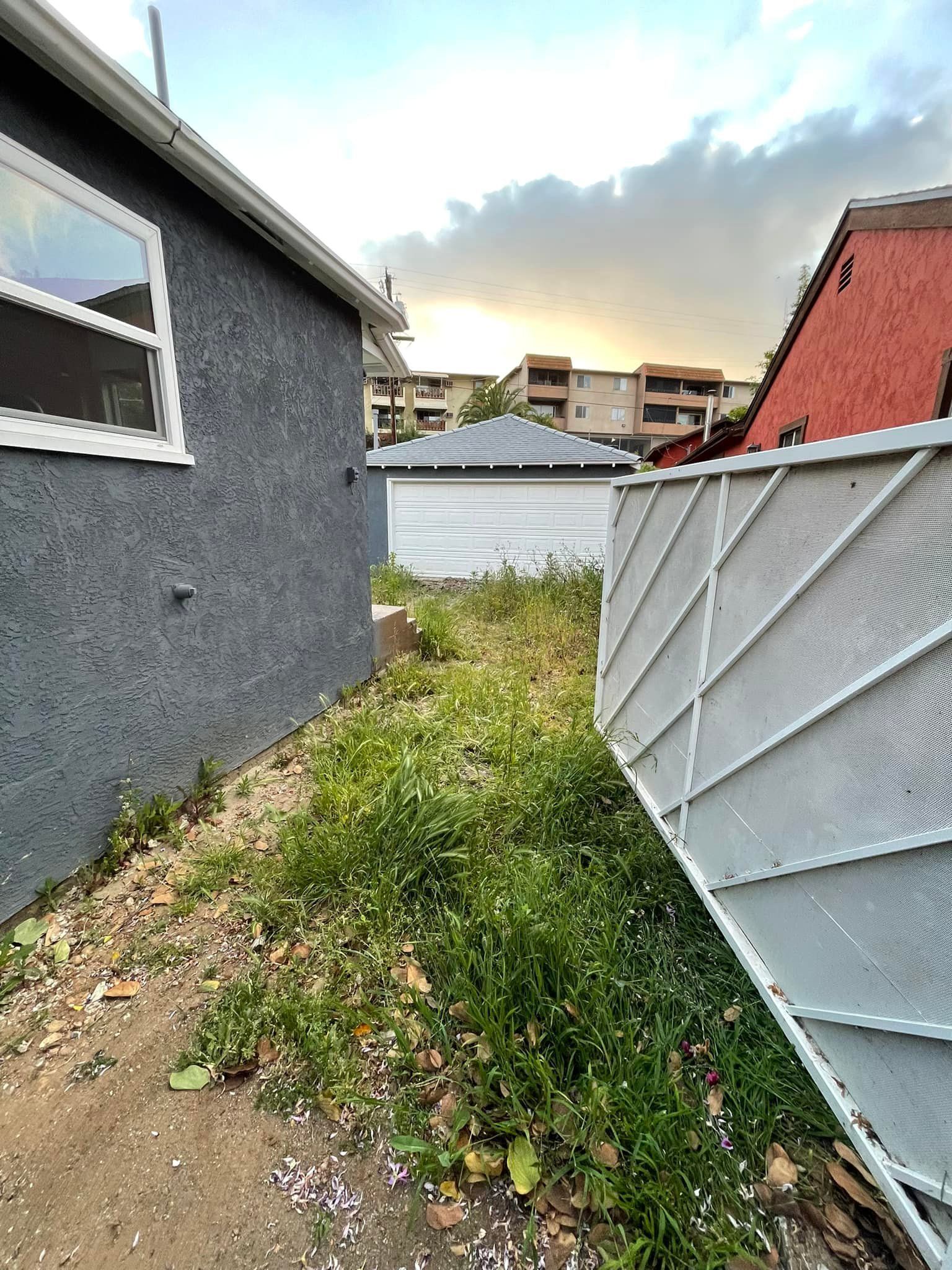 Overgrown grassy alley between gray stucco building and white slatted fence, with a garage and buildings in the background.
