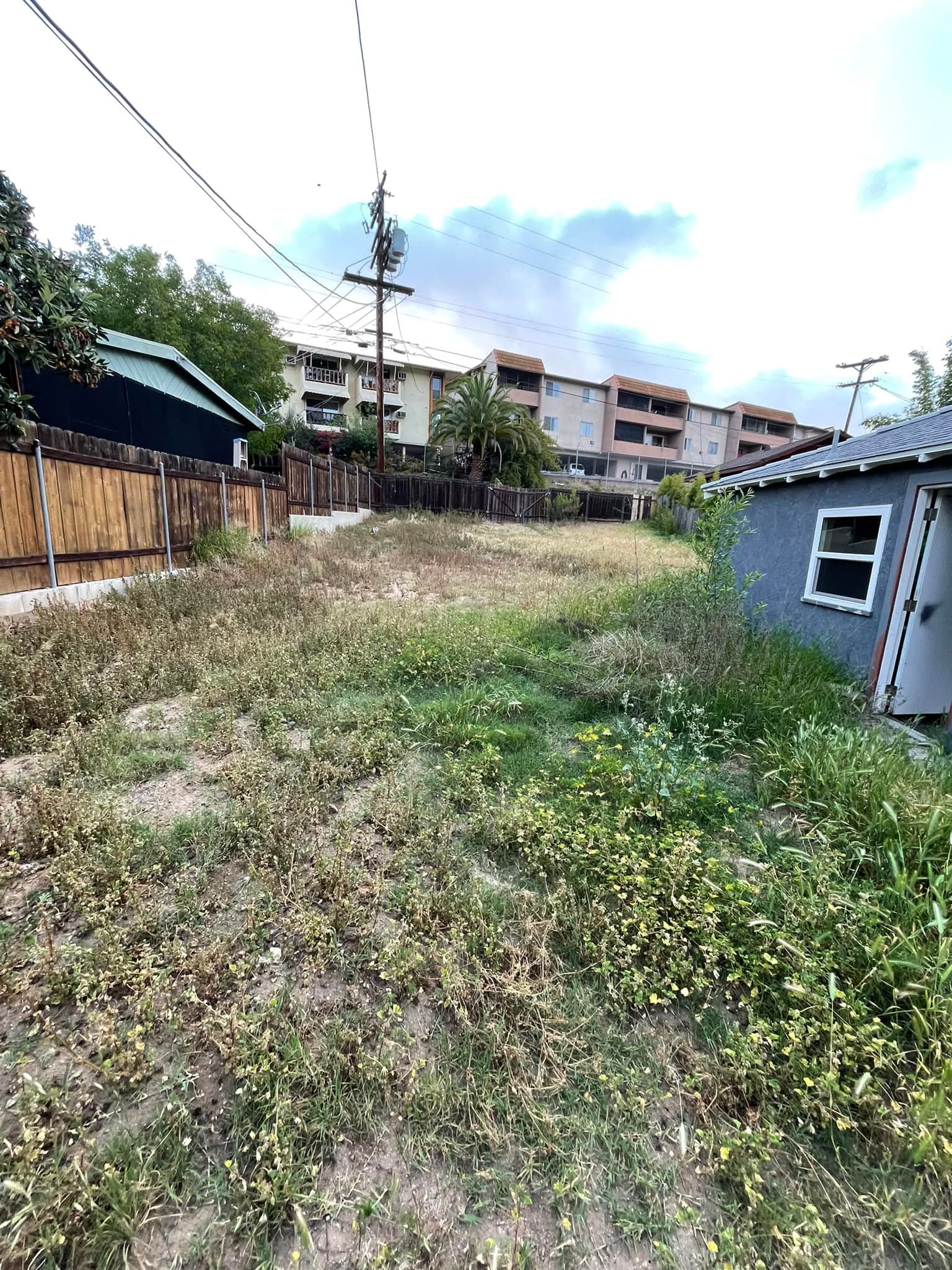 Grassy lot with weeds, bordered by fences and buildings under a cloudy sky.