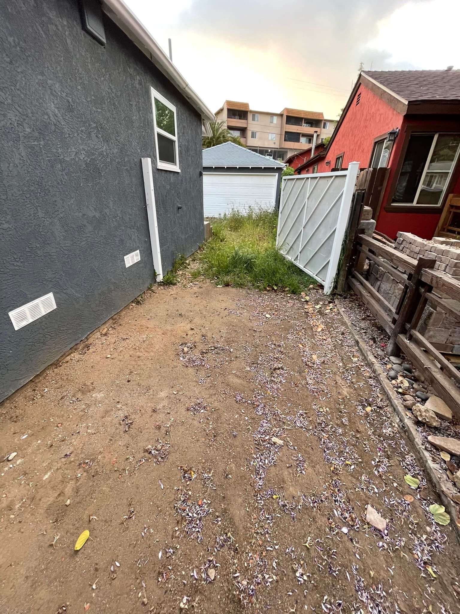 Narrow dirt path between a gray building and a white fence with overgrown weeds, and a red building in the distance.