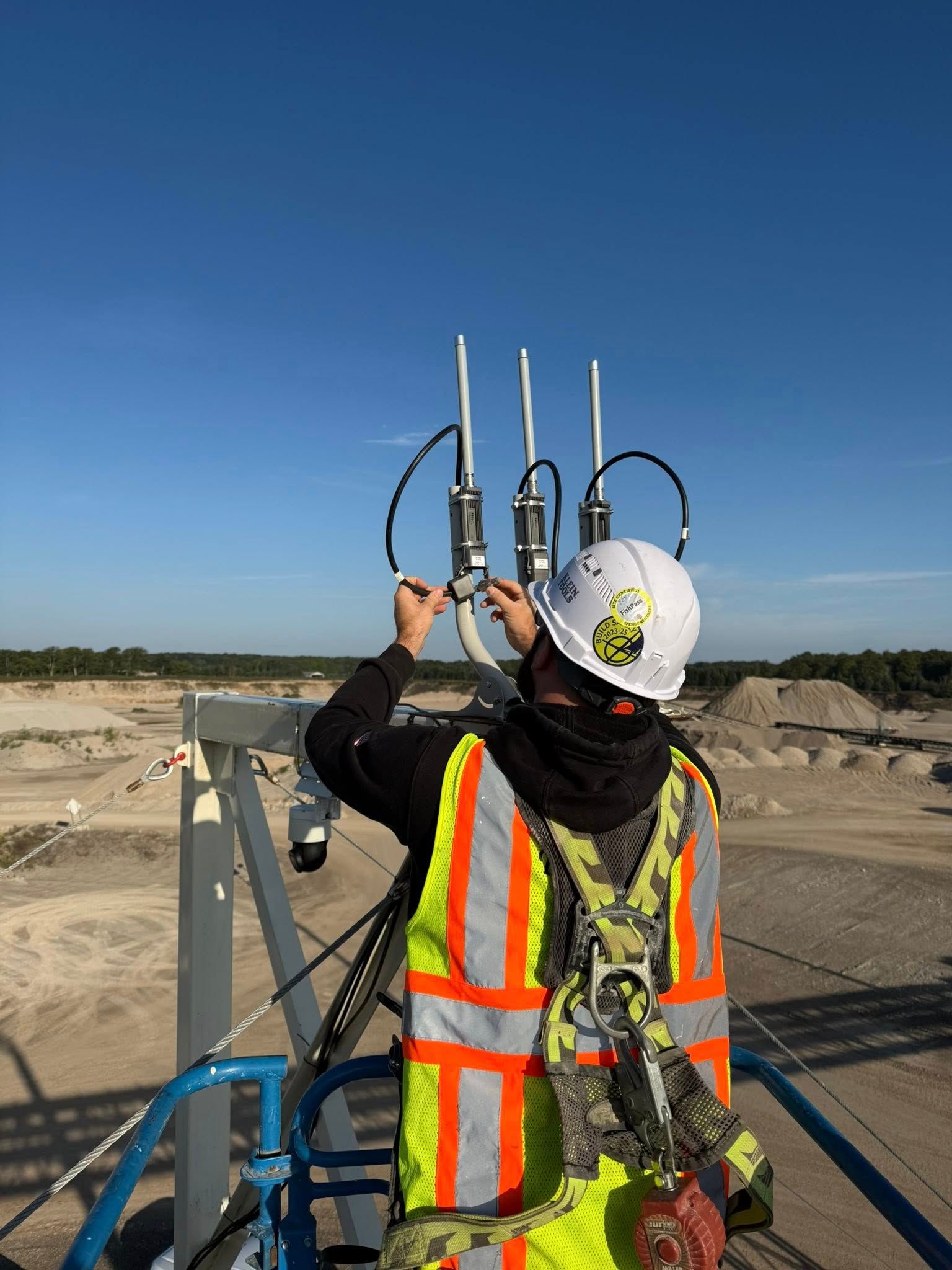 Worker in safety vest on a lift adjusts equipment atop a structure at a construction site against a blue sky.