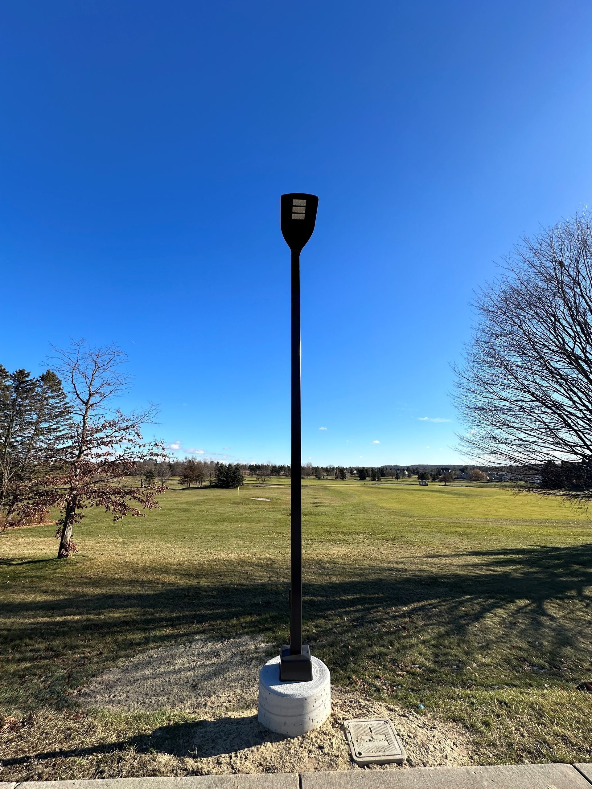 Black lamppost against a clear blue sky, concrete base on a grassy field.