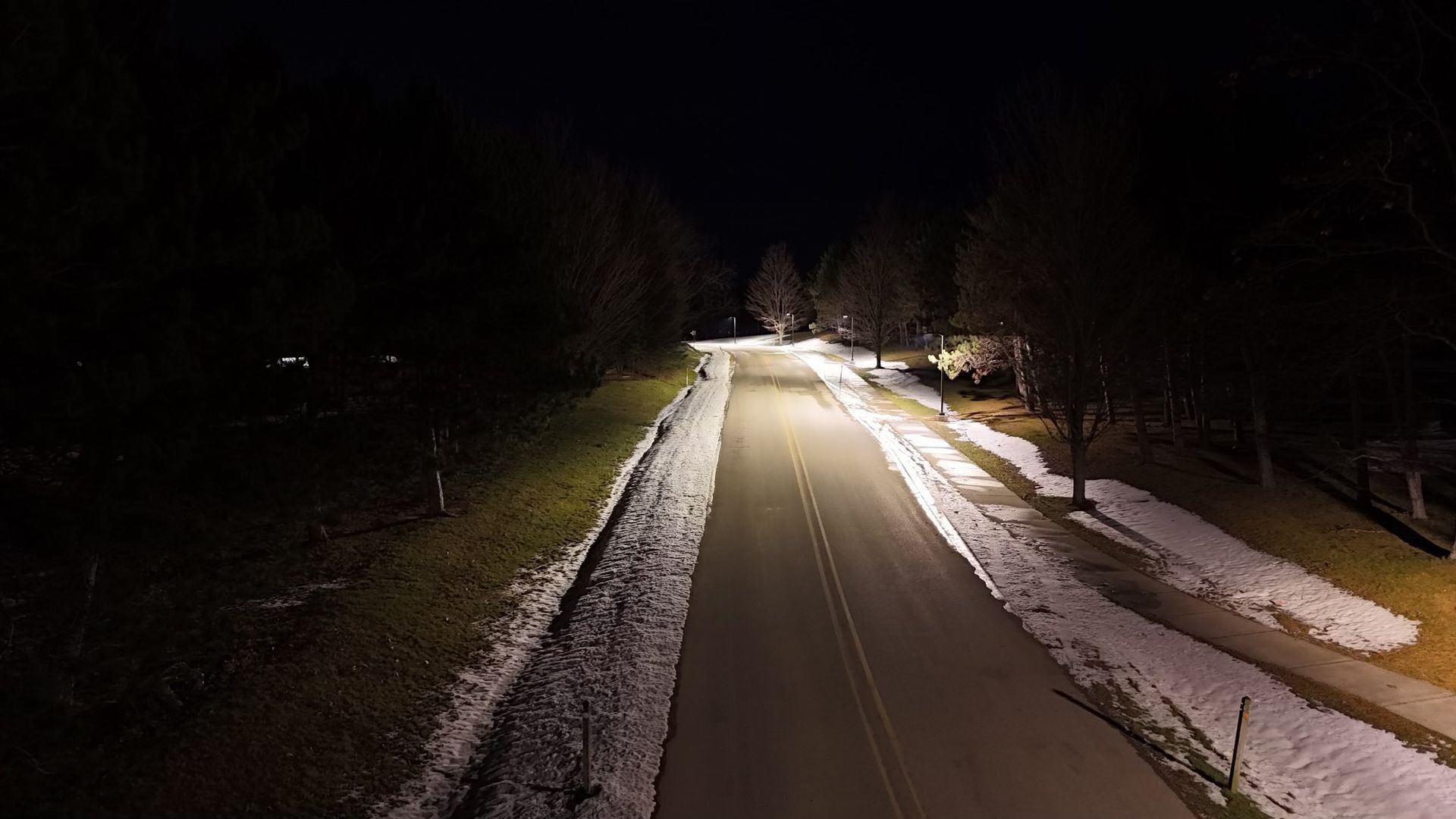 Nighttime aerial view of a road with snow-covered shoulders, flanked by trees and illuminated by streetlights.