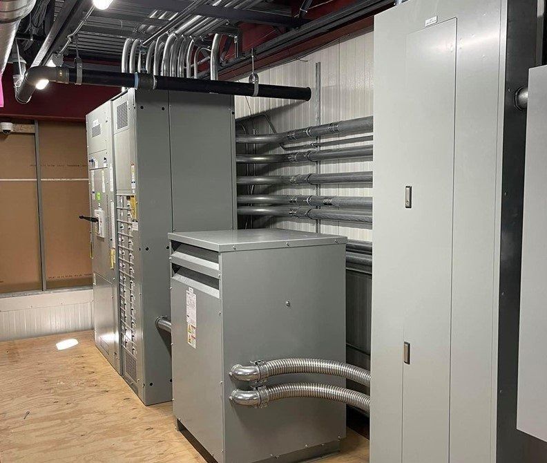Electrical equipment room with gray metal panels, conduit, and ductwork on a light wood floor.