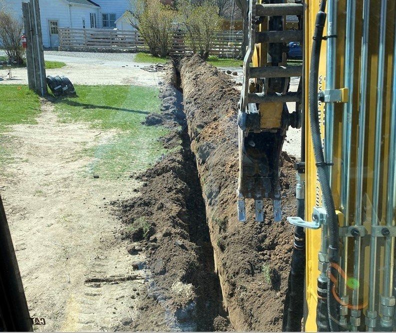 A yellow excavator digging a trench in the dirt, near a grassy area and a gravel driveway.