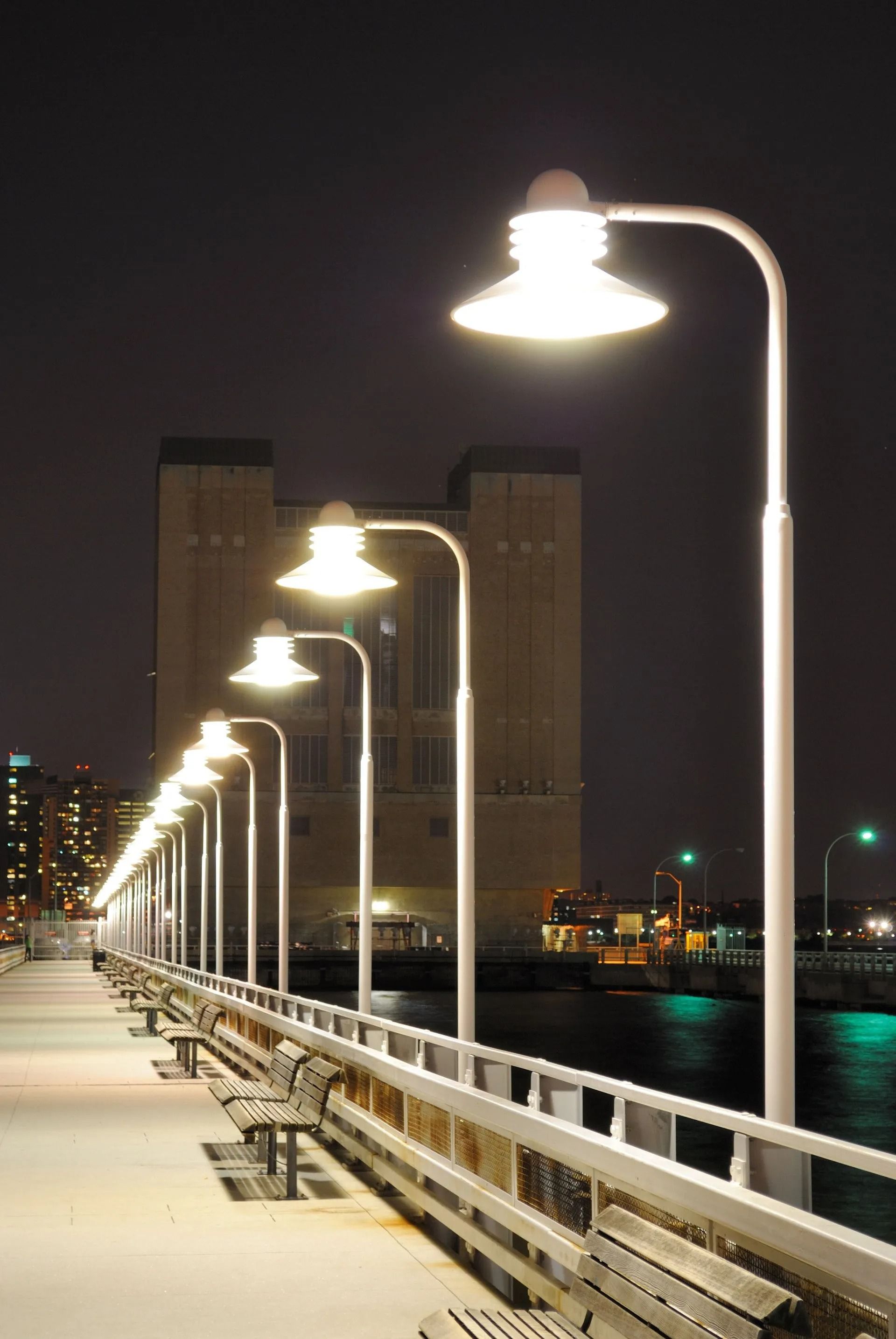 Night view of a snowy pier lined with glowing streetlights, overlooking dark water and a distant building.