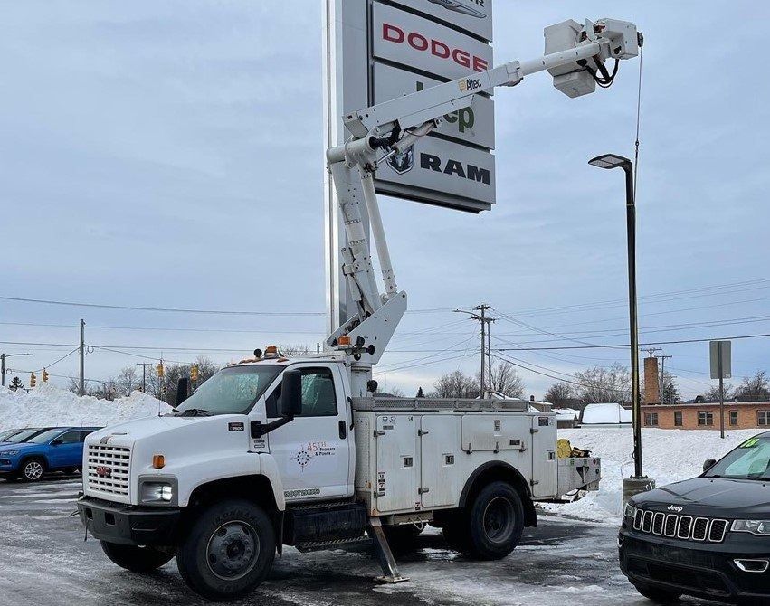 Truck-mounted lift servicing a Dodge RAM sign at a dealership, with a white and cloudy sky backdrop.