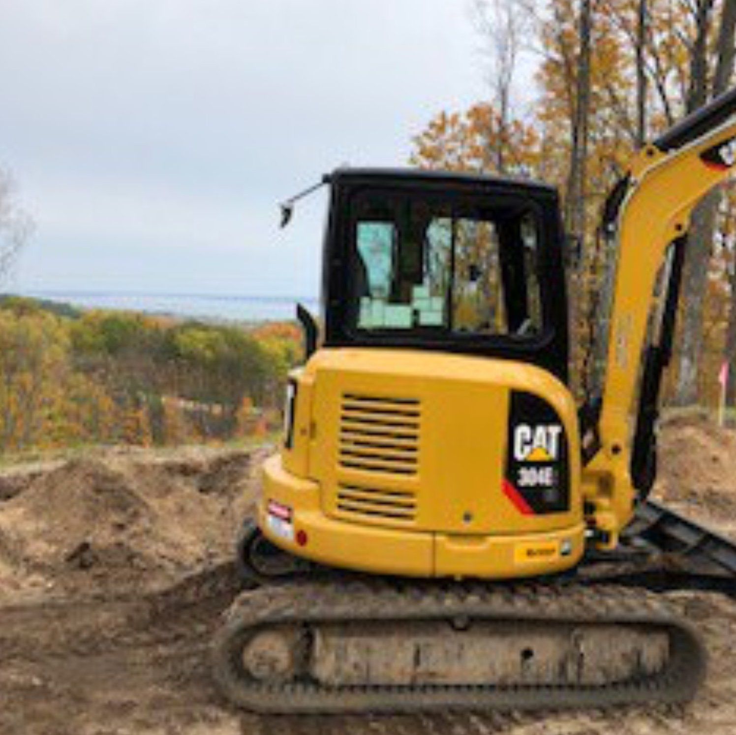 Yellow Caterpillar 304E mini excavator on a construction site, with a forest and body of water in the background.