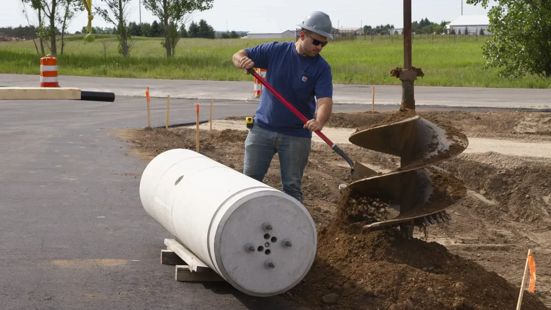 Man using an auger to dig a hole next to a concrete cylinder on a construction site.