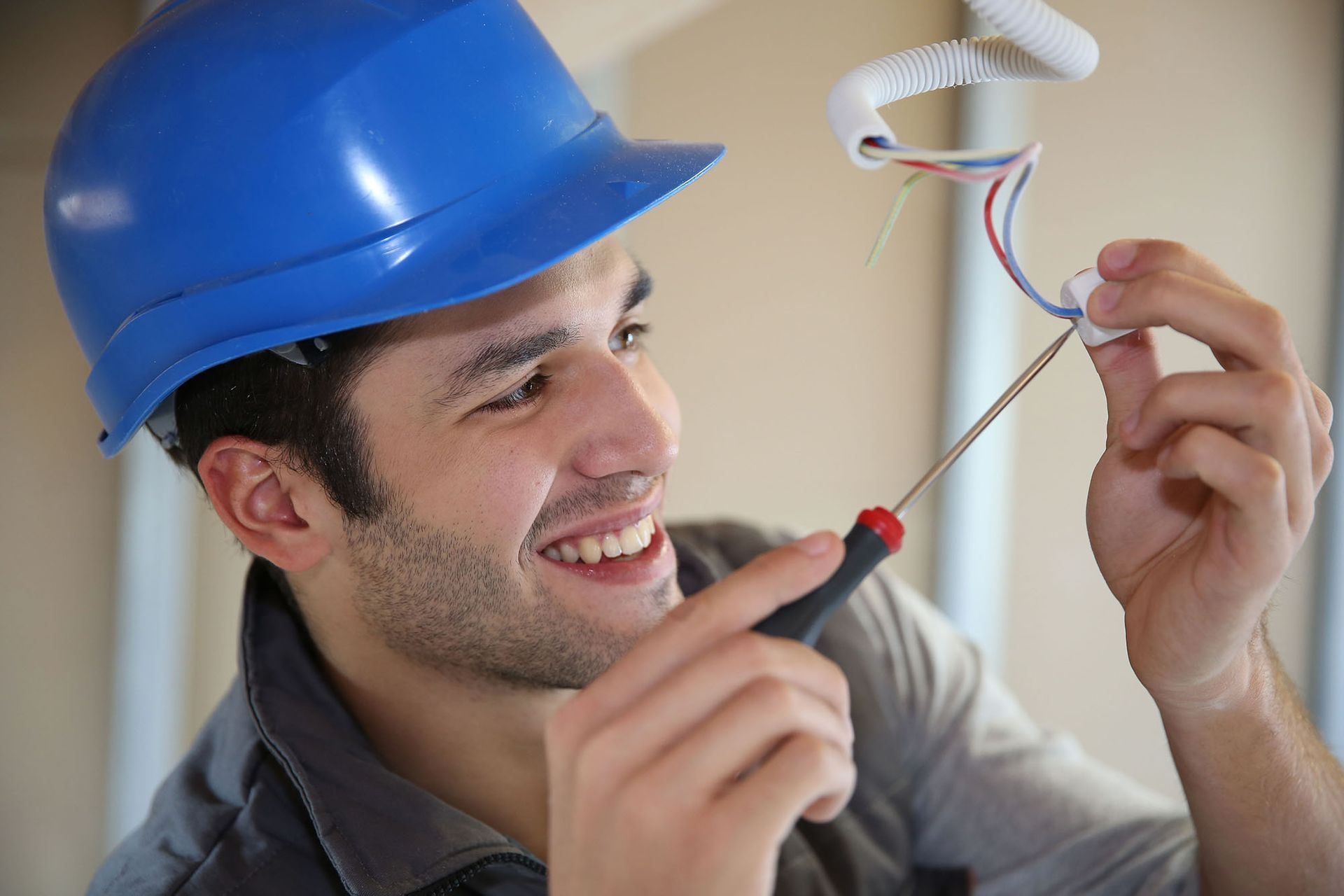 Electrician in a blue hard hat smiles while wiring.